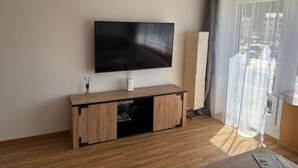Un salon moderne avec un téléviseur à écran plat fixé au mur, une console en bois intégrant un meuble pour appareils multimédias, et une grande fenêtre ornée de rideaux légers.