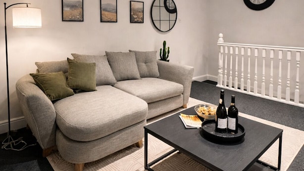 Una sala de estar moderna con un sofá gris, una mesa de centro negra con una botella, arte enmarcados, un reloj y una barandilla blanca.