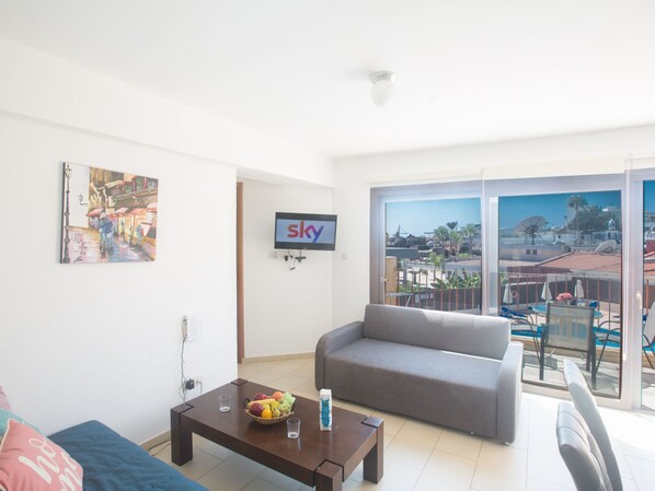 Ein Wohnzimmer mit einer Couch, einem Couchtisch mit Früchten, einem Fernseher an der Wand und einem Balkon mit Blick auf die Stadt.
