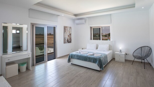 A modern bedroom with a bed, bedside tables, a chair, and a view through a sliding door. - Geräumiges Ferienhaus für 12 Personen in Paralini by Interhome (Paralimni)