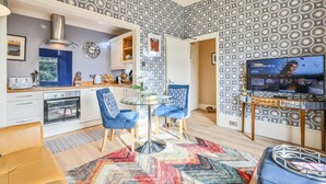 Une cuisine moderne avec des armoires blanches, un four encastré et une fenêtre. On y trouve un coin repas avec des chaises bleues et une table en verre. Un téléviseur est fixé au mur et diffuse une émission.