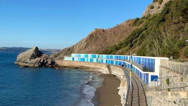 Une promenade en bord de mer, agrémentée d’une barrière en pierre et de panneaux bleus, qui mène à une plage parsemée de rochers et offrant une vue dégagée sur la mer.