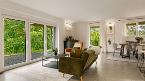 Amplia sala de estar con sofá, mesa de centro y mesa de comedor. Grandes ventanales ofrecen vistas a la vegetación exterior.