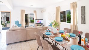 Een moderne woonkamer met een bank, een eettafel gedekt met blauwe borden en glazen, een televisie waarop 'lucht' staat en een balkon met gordijnen.