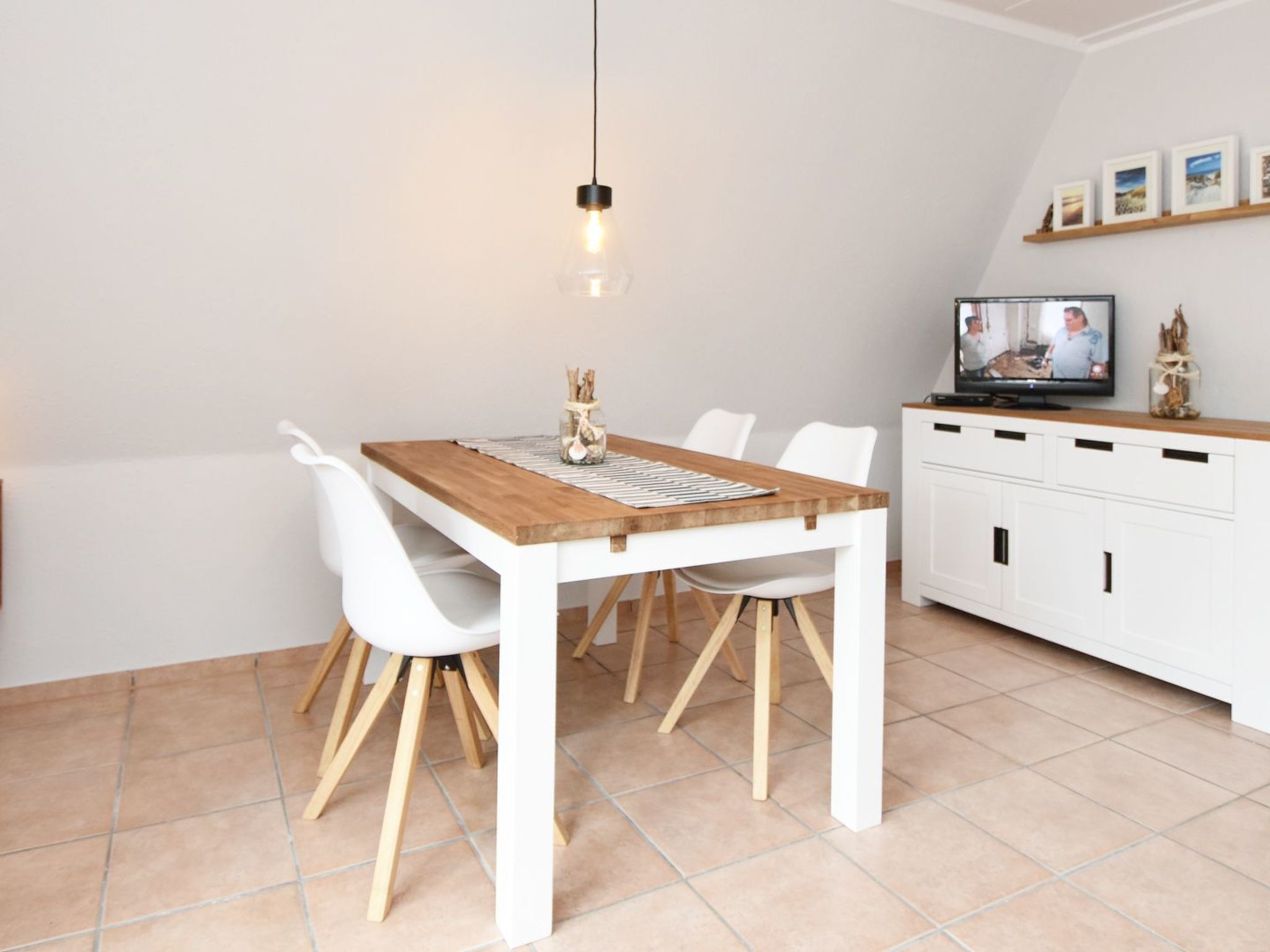 Una cocina moderna con una mesa de madera, sillas blancas, una lámpara colgante, un televisor y un armario con repisa superior.