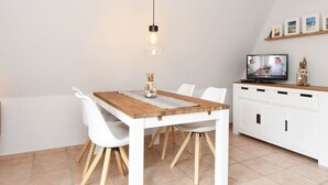 A modern kitchen with a wooden table, white chairs, a hanging light, a TV, and a cabinet with a shelf above. - Carolin by Interhome (Baabe)