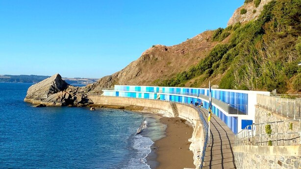 Een kustpad met een stenen muur en blauwe leuningen, dat naar een strand leidt met helderblauw water en een rotsachtige klif.