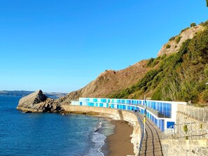 Ein Küstenweg mit einer Steinmauer und blauen Geländern, der zu einem Strand mit klarem, blauem Wasser und einer felsigen Klippe führt.