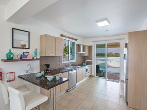 A modern kitchen with a central island, a dining area with a white chair, and a sliding glass door leading to a balcony with a pool. - 3-Schlafzimmer-Villa mit privatem Pool by Interhome (Agios Georgios Pegeias)