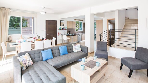 A modern living room with a grey sectional sofa, a coffee table, and a grey armchair. There is a dining area with a table and chairs, and a kitchen in the background. - 3-Schlafzimmer-Villa mit privatem Pool by Interhome (Agios Georgios Pegeias)