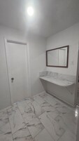 Un baño con puerta blanca, suelo de mármol, tocador blanco con espejo y una lámpara en el techo.