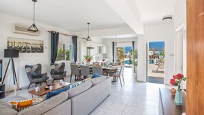 Ein großzügiger Wohnbereich mit einer großen Couch, einem Esstisch und Stühlen. Im Hintergrund befindet sich eine Küchenzeile, und es gibt einen Blick auf den Pool und eine Außenlounge.