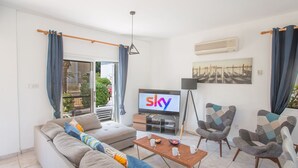 Ein modernes Wohnzimmer mit einer Couch, Sesseln, einem Couchtisch, einem Fernseher und einem Balkon mit Vorhängen.