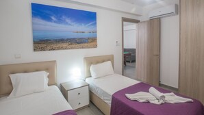Hotellihuone, jossa on kaksi sänkyä, yöpöytä, vaatekaappi ja taulu, jossa on kuvattuna rantamaisema.