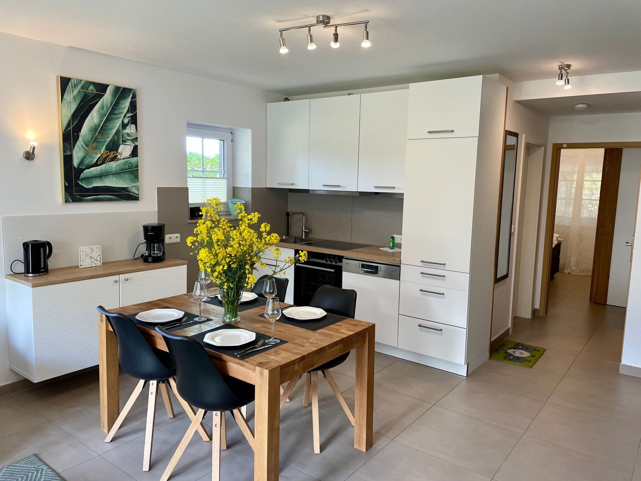 A modern kitchen with white cabinets, a wooden dining table, and a vase of yellow flowers.