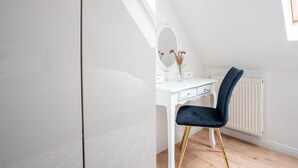 A white vanity with a mirror, a vase with flowers, and a dark blue chair with gold legs in a room with wooden flooring and a white radiator. - Schule Altes Land by Interhome (Jork)