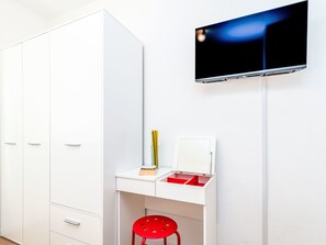 Ein Zimmer mit einem weißen Kleiderschrank, einem Schreibtisch mit einem roten Stuhl, einem an der Wand befestigten Flachbildfernseher und einer Vase auf dem Schreibtisch.
