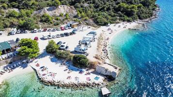 An aerial view of a coastal area with a beach, clear blue waters, and a row of RVs parked on the sand.