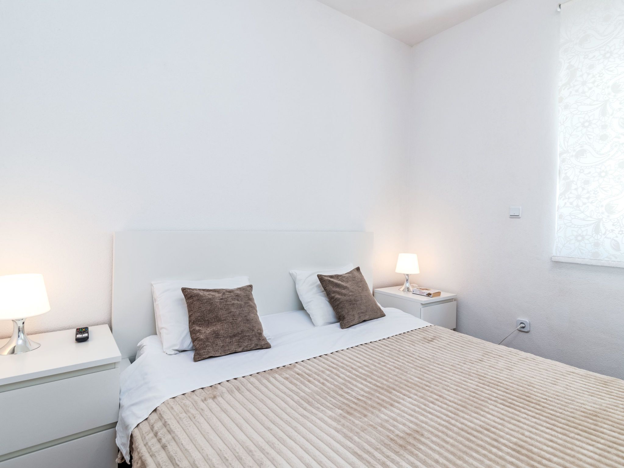 Habitación de hotel minimalista con cama, dos lámparas de noche, una mesita de noche y un ventanal con cortina estampada.