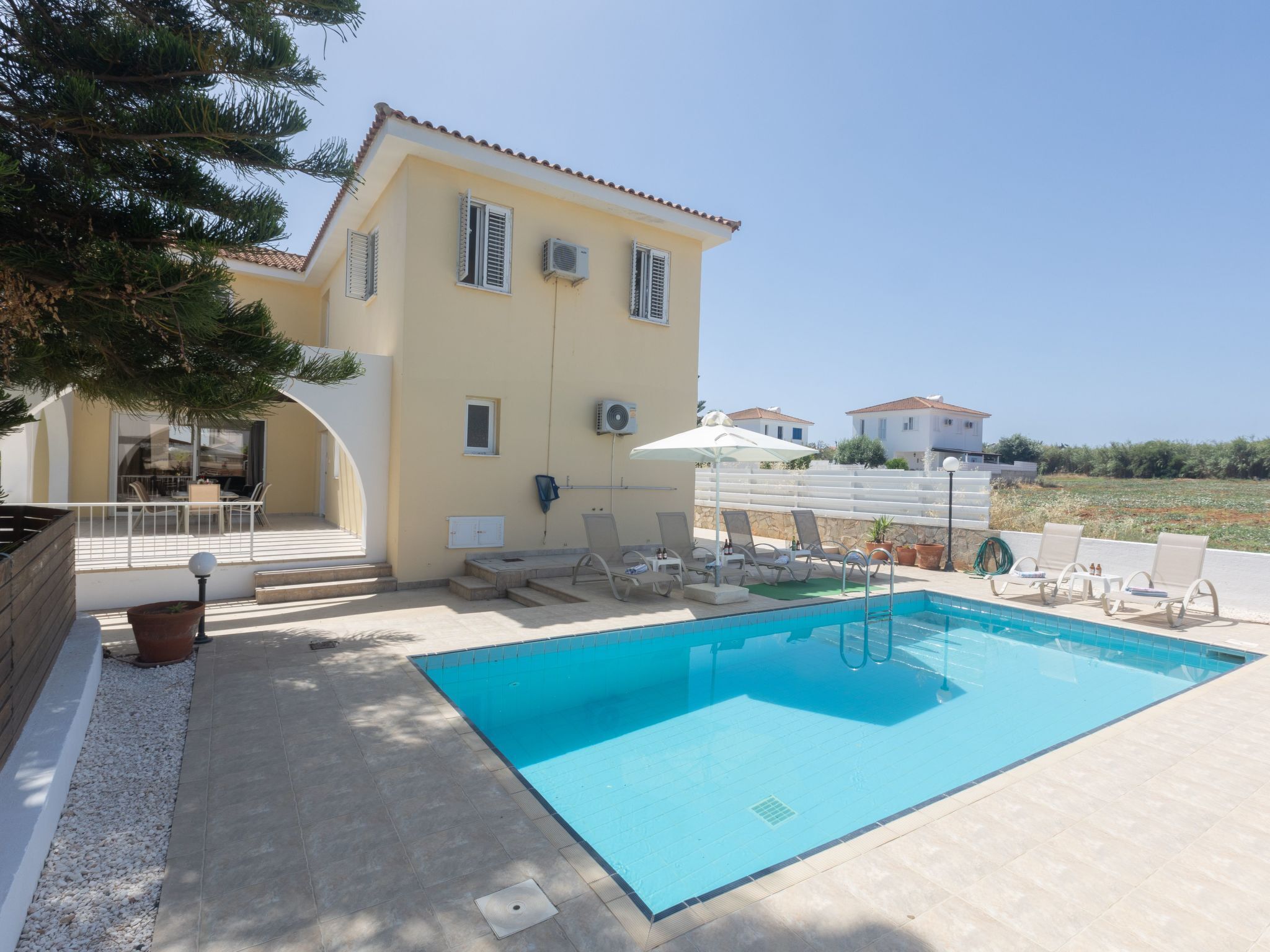 A pool area with a clear blue pool, sun loungers, and a building with a tiled roof and shutters.