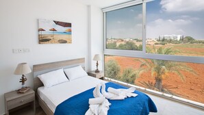 Een hotelkamer met een groot raam, een bed met een blauwe sprei, een nachtkastje met een lamp, en een schilderij van een strand.