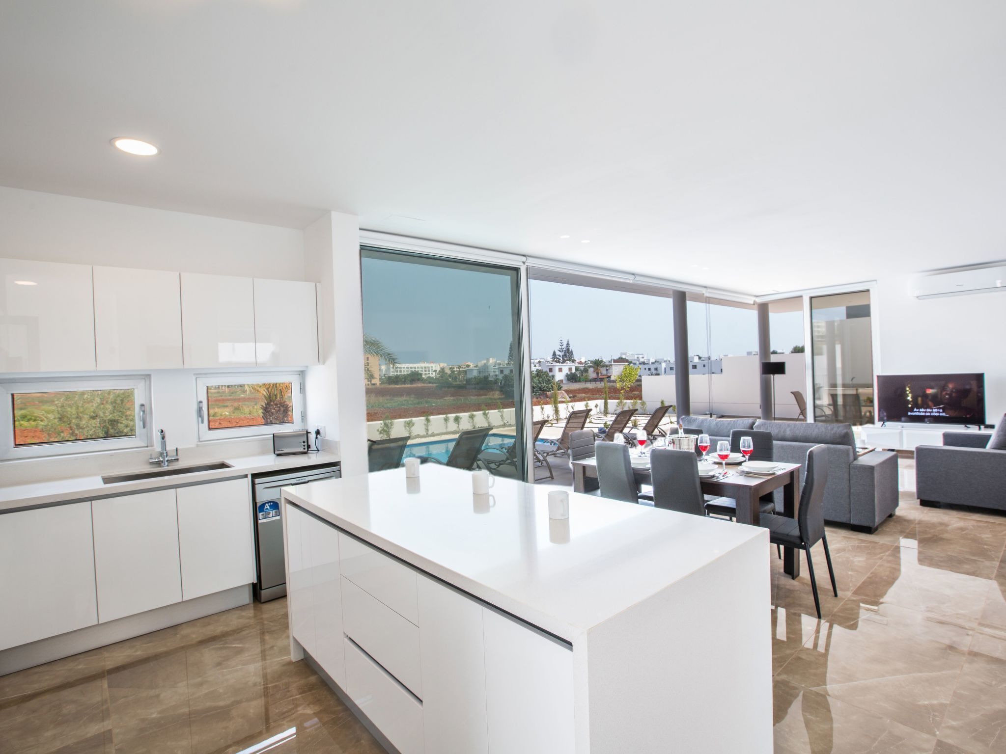 A modern kitchen with white cabinets and a large island, a dining area with a pool view, and a living room with a flat-screen TV.