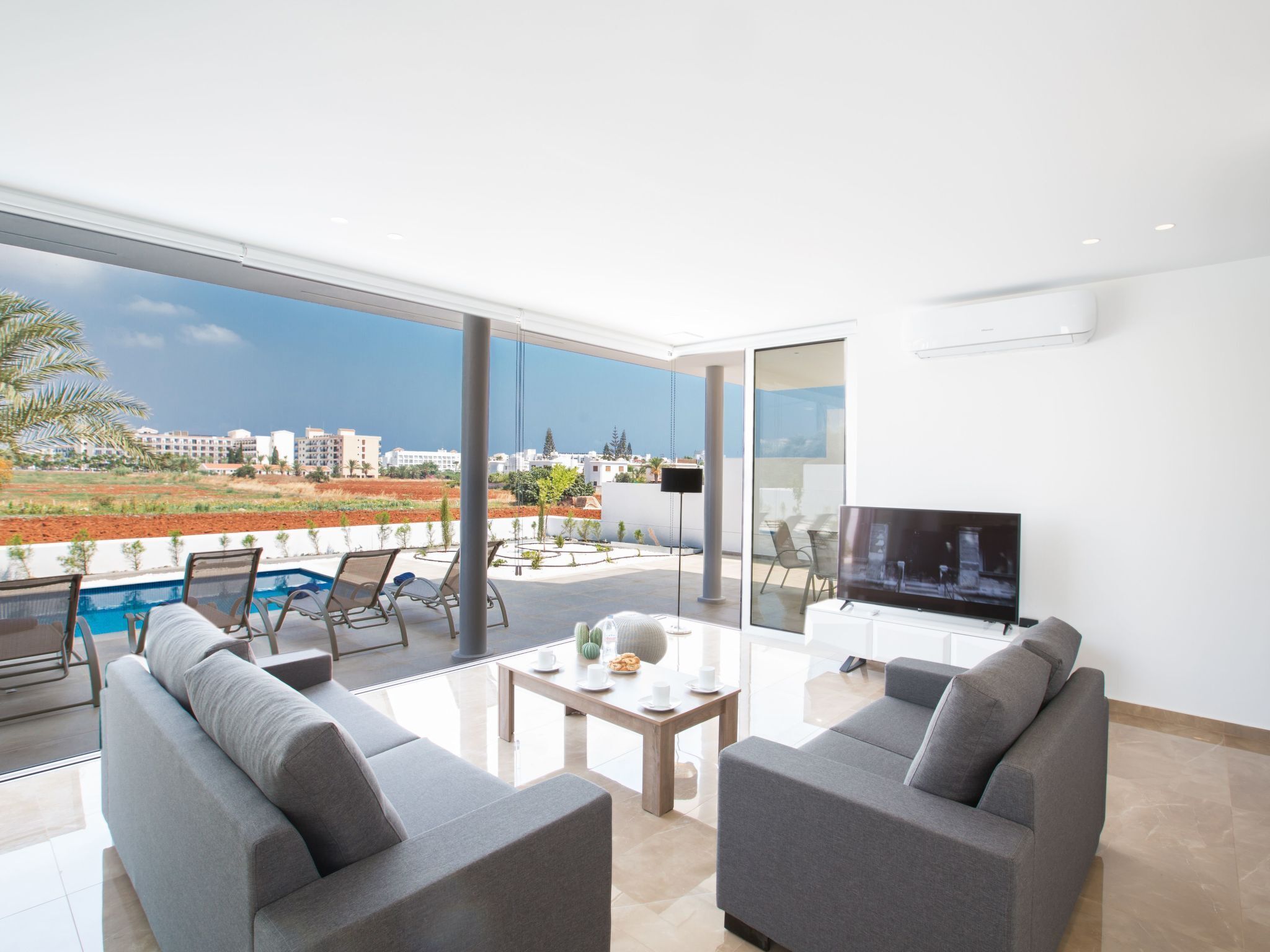 A modern living room with a large TV, grey sofas, a coffee table, and a pool view.