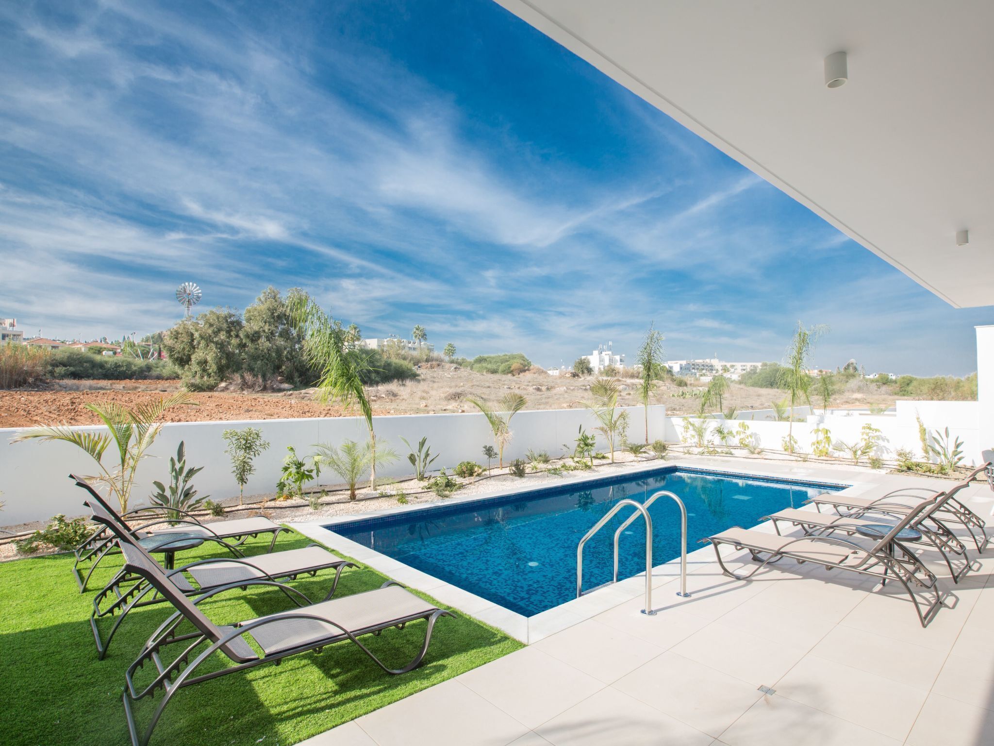 A modern outdoor pool area with a clear blue sky, surrounded by greenery and white walls.