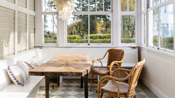 Ein Zimmer mit großem Fenster, einem Holztisch, Korbstühlen und einer Bank mit Kissen.