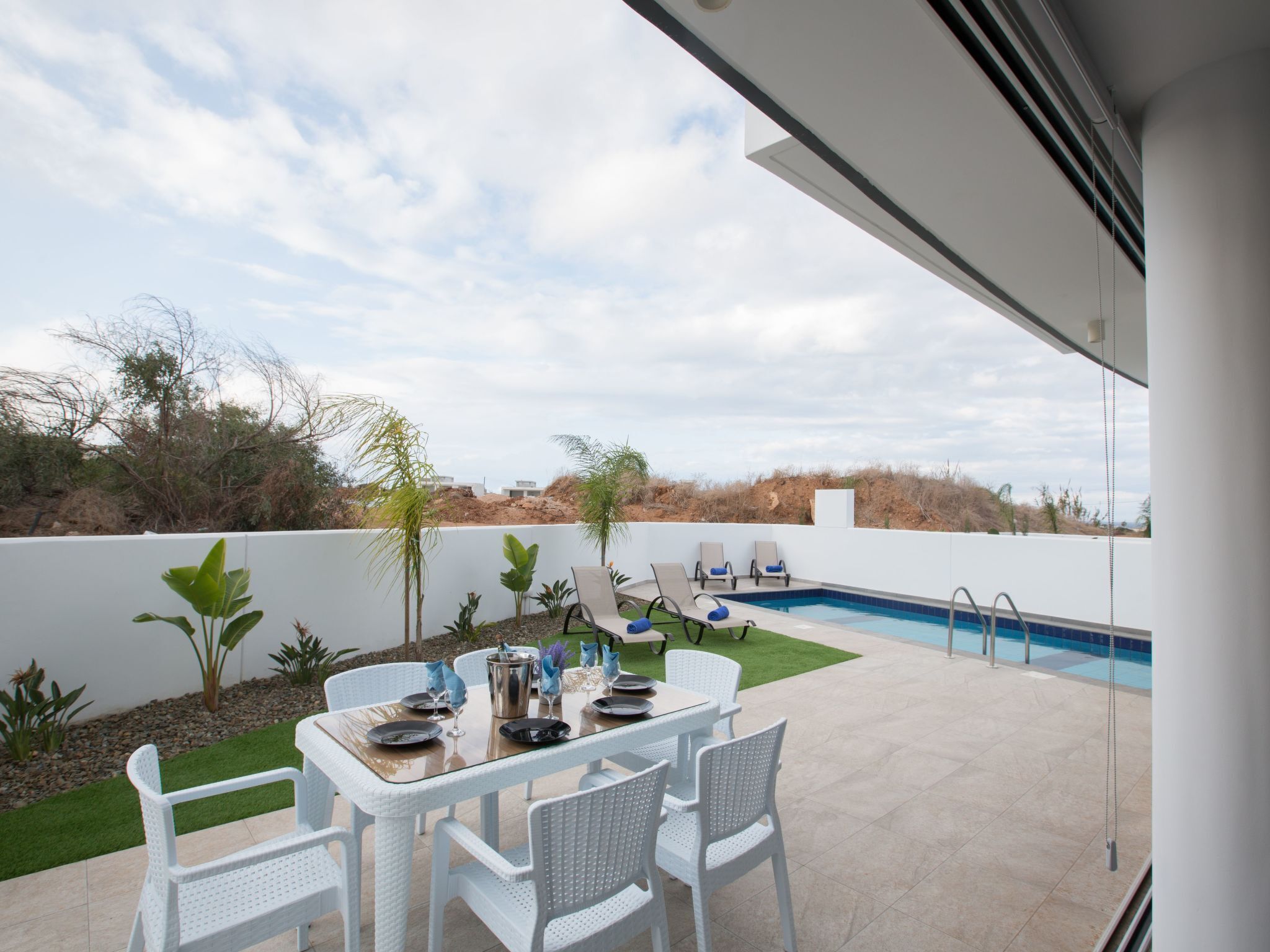 Zona junto a la piscina con una mesa dispuesta para comer, rodeada de sillas blancas, y una piscina con agua cristalina.