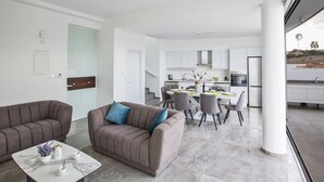 Una sala moderna con sofá y mesa de centro, un comedor con sillas y mesa, y una cocina con gabinetes blancos y ventana.