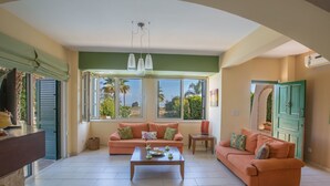Uma sala de estar espaçosa com dois sofás, uma mesa de centro e vista para palmeiras através de grandes janelas.