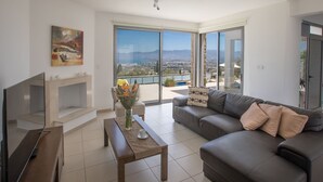 A modern living room with a large flat-screen TV, a wooden coffee table, a grey sofa, and a view of a cityscape through large sliding glass doors. - Villa mit Panoramablick auf das Meer von Latchi by Interhome (Polis Chrysochous)