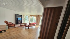 Uma sala de estar ampla com piso de madeira, um sofá, uma poltrona e uma televisão.