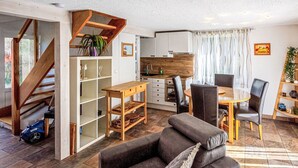 Un moderno salón con sofá, mesa de comedor y sillas. Se ve una escalera que conduce a otro nivel, una zona de cocina y un ventanal con cortinas.
