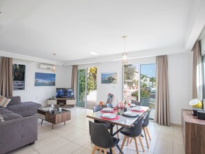 Ein modernes Wohnzimmer mit Sofa, Couchtisch, Fernseher und Essbereich mit Stühlen und gedecktem Tisch.