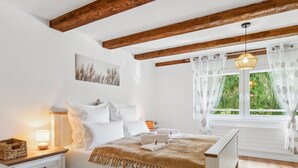Ein Schlafzimmer mit Holzboden, einem Bett mit weißer Bettwäsche, einem Nachttisch mit Lampe und einem Fenster mit Vorhängen.