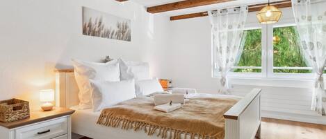 Une chambre avec un plafond en bois, un lit recouvert de linge de lit blanc, une table de chevet avec une lampe et une fenêtre avec des rideaux.