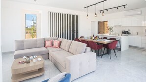 A modern living room with a sofa, coffee table, and dining area with chairs. There is a kitchen in the background with white cabinets and a dining table. - Protaras Vie Bleu Villa mit Pool, im Zentrum by Interhome (Protaras)