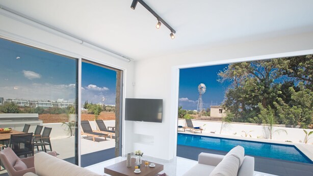 Ein modernes Wohnzimmer mit einem großen Glasfenster zum Poolbereich, einem wandmontierten Flachbildfernseher und Blick auf eine Windmühle und grüne Natur.
