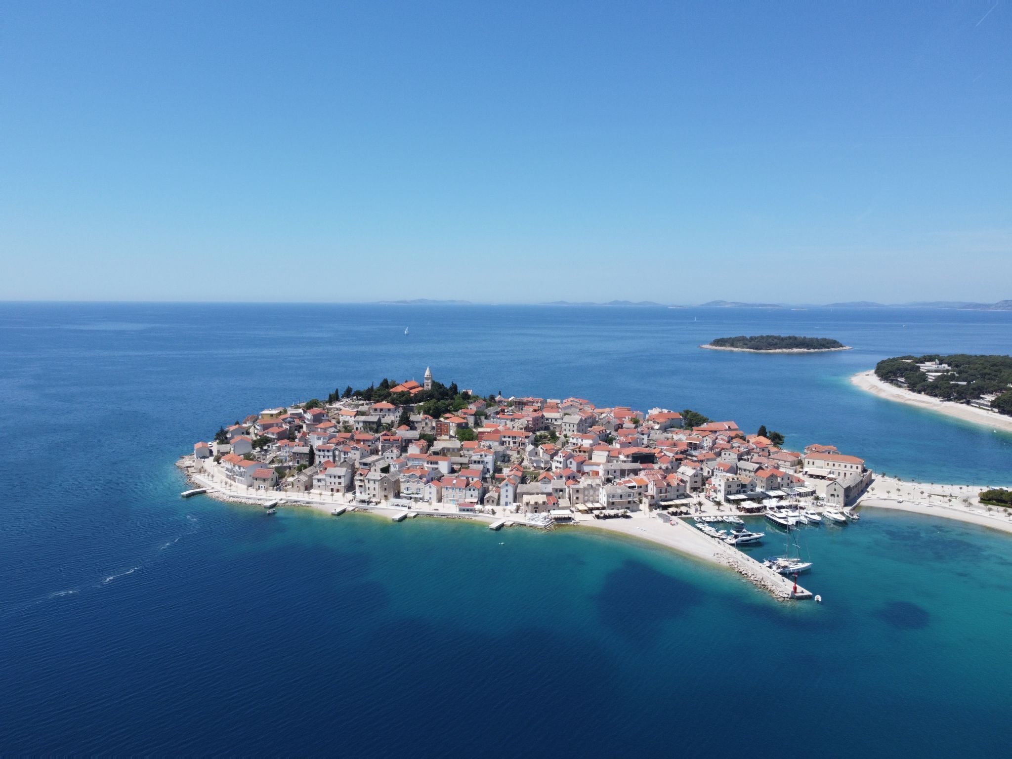 Vista aerea di una cittadina costiera con edifici raggruppati attorno a un’area centrale, collegati alla terraferma da una stretta lingua di terra.