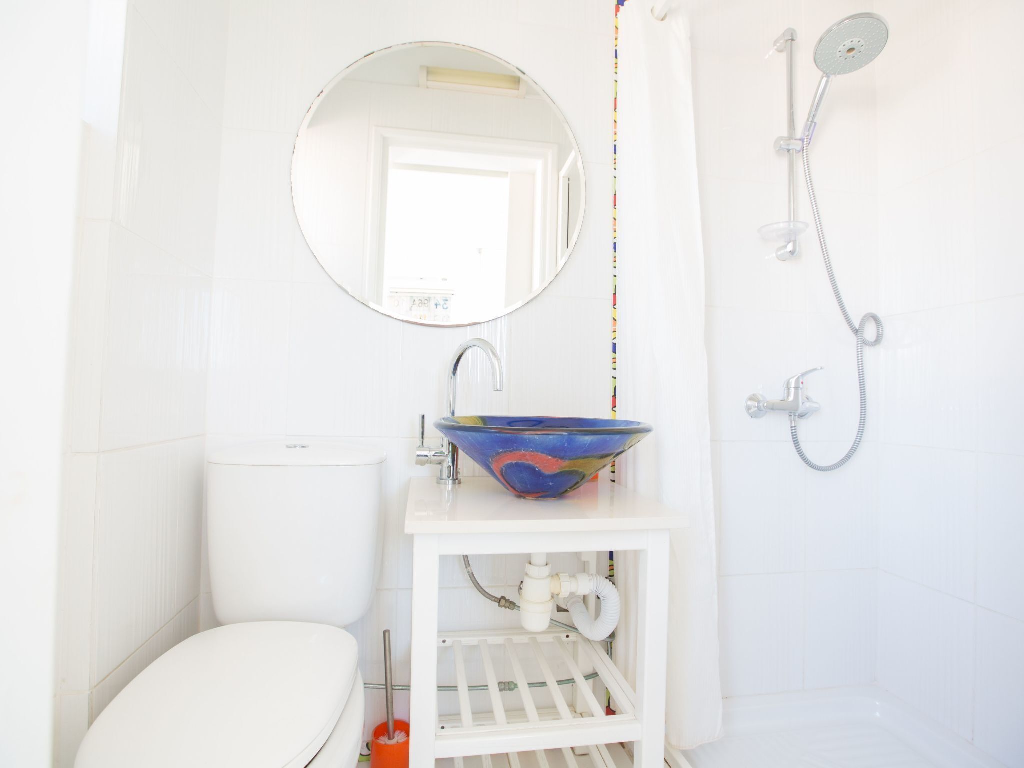 A bathroom with a white toilet, a round mirror, a blue bowl sink, and a shower.