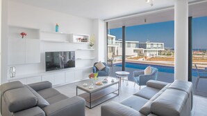 A modern living room with a flat-screen TV, grey sofas, a coffee table, and a view of a swimming pool and buildings. - Schöne Villa Olivine Pearl OL5 mit Garten by Interhome (Protaras)