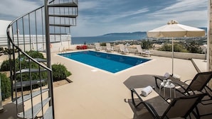 Une piscine sur le toit offrant une vue dégagée sur la mer et les montagnes, un escalier en colimaçon et un espace extérieur aménagé avec des parasols.