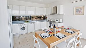 Une cuisine moderne avec des meubles blancs, un lave-vaisselle et un coin repas comprenant une table en bois et des chaises blanches.