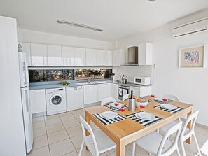 A modern kitchen with white cabinets, a dishwasher, and a dining area with a wooden table and white chairs. - Latchi Ferienvilla EV20 by Interhome (Agios Georgios Pegeias)