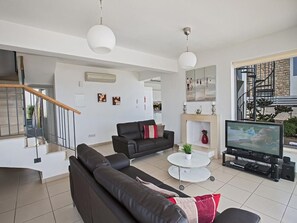 A modern living room with a black leather sofa, a white coffee table, and a TV. There is a staircase leading to another level, and a fireplace with a mantel. - Latchi Ferienvilla EV20 by Interhome (Agios Georgios Pegeias)