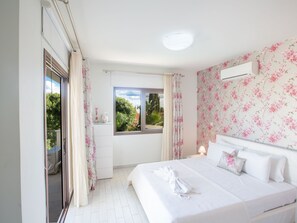 Um quarto com cama, janela com cortinas, mesa de cabeceira e ar-condicionado fixado na parede.