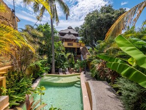 Un área de piscina con un edificio de techo de paja, rodeado de exuberante vegetación tropical.