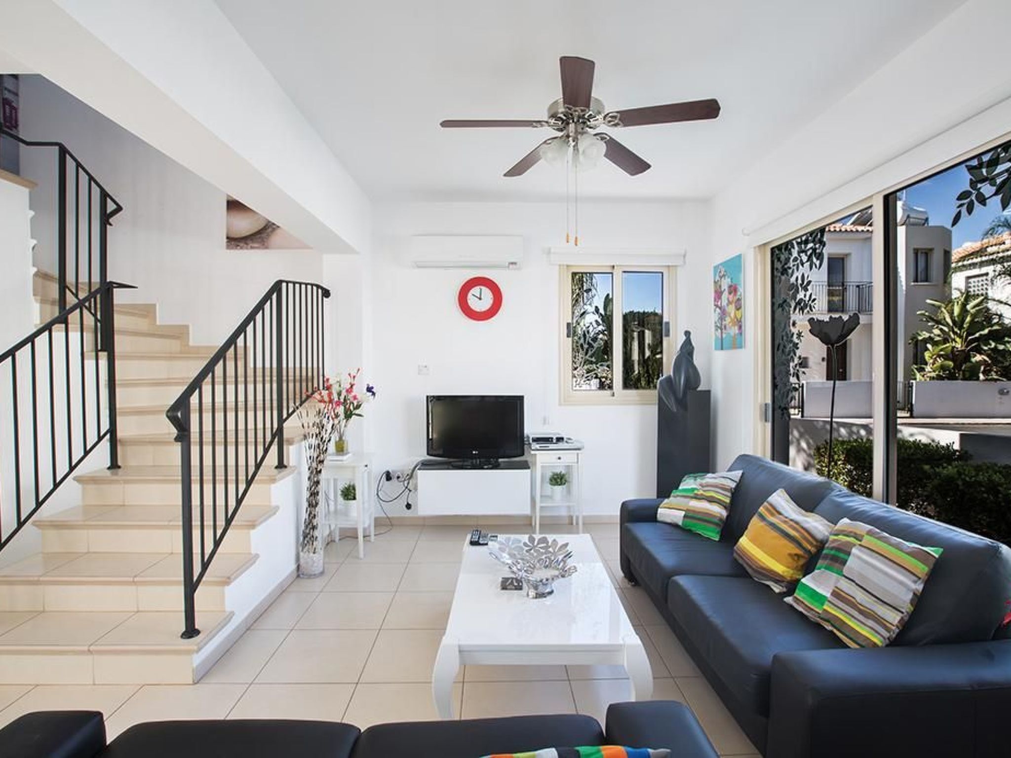 A modern living room with a staircase, a ceiling fan, a flat-screen TV, a coffee table, and a sofa with colorful cushions.